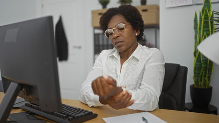 Woman working on a computer in an office while holding her wrist, indicating discomfort, with curly hair and glasses, surrounded by indoor plants and office decor in the background