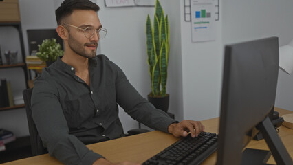Handsome young hispanic man with glasses and beard working on a computer in a modern office setting featuring plants and shelves in the background.
