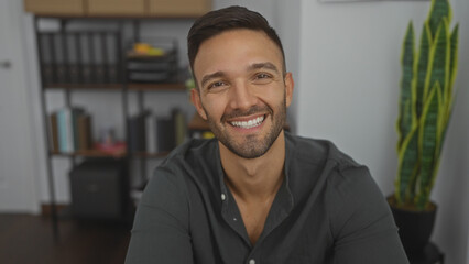Fototapeta premium Handsome young hispanic man with a beard smiling in an indoor office setting with shelves and a plant in the background.
