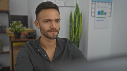 A handsome young hispanic man with a beard is focused at his desk in an indoor office setting, surrounded by plants and documents on the wall.