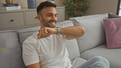 Hispanic man with a beard looking at his watch and smiling while sitting in a cozy living room at home.