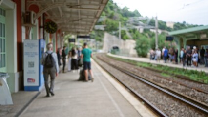 People wait at train station with blurred background highlighting defocused urban setting bustling with travelers and commuters on railway platform.