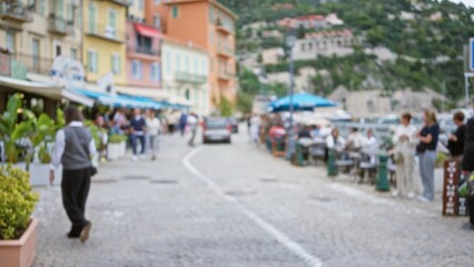 Blurred view of people walking in sunny villefranche-sur-mer, france, featuring colorful buildings and outdoor cafes along a cobblestone street with vibrant foliage.