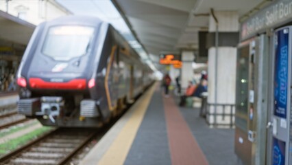 Blurred train moves through defocused railway station with bokeh lights and travelers waiting on platform in bustling urban scene