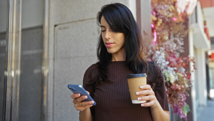 Young woman in urban street holding coffee and smartphone, showing a hispanic adult female in an outdoor city setting