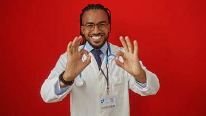 Handsome young black man with braids makes an ok gesture against a vibrant red wall, exuding positivity and confidence, dressed in a doctor or professional outfit.