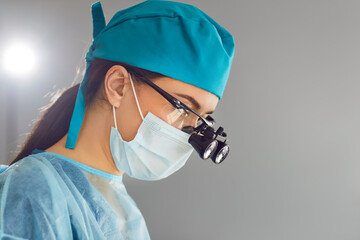 Qualified focused female surgeons wear magnifying glasses in operating room. Close-up side view of young woman wearing medical protective mask, surgical cap and intraoperative magnifying glasses.