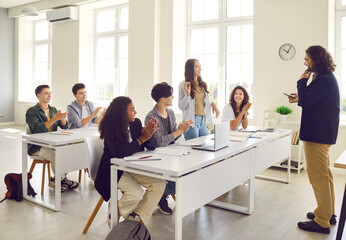 Young girl student standing at the desk in classroom and answering the male teacher with a college or high school students applauding her during a lesson. Education and learning concept.