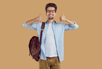 Happy university student guy with leather backpack showing thumbs up on beige background. Happy...