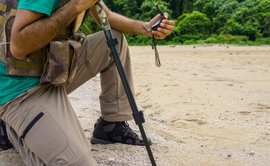 Man kneels with a compass and hiking stick on a sandy beach, focused on navigation and exploration.