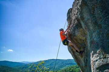 Aerial view of male rock climber ascending rugged limestone cliff with harness and rope for safety....