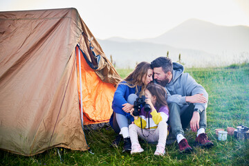 Family of three sits together in front of orange tent in grassy mountain campsite. Child uses...