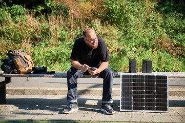Bearded man using smartphone connected to photovoltaic solar panel. Integration of sustainable...