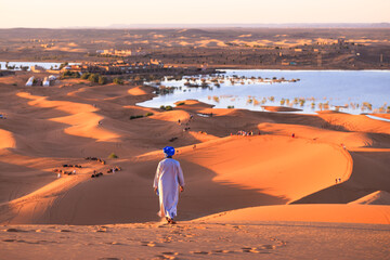 A tuareg in traditional clothing, hiking through the dune landscape of Erg Chebbi with lake Yasmina...