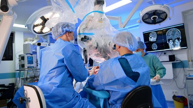 Cooperating surgeons perform operation and look at microscope. Monitor displaying brain MRI scan at backdrop. Neurosurgery concept.