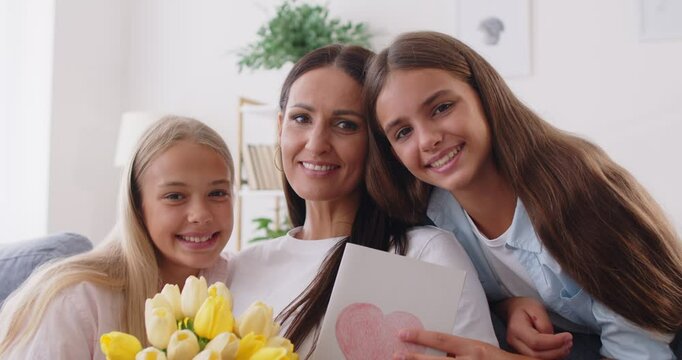 Portrait of happy mother and two preteen daughters, embracing in living room, celebrating Mother Day, birthday or Women Day, making family photo with flowers and card, looking at camera and smiling.