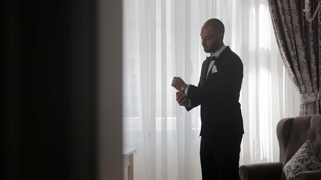 Groom standing by window adjusting cufflinks, A groom in a sleek black tuxedo stands by a sunlit window, adjusting his cufflinks