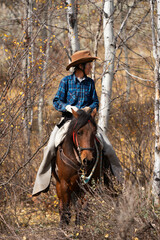 Fototapeta premium elderly woman in western hat and attire riding a bay Morgan mare in the fall aspen leaves in the mountains of Wyoming