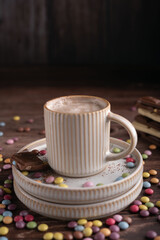 photo Cozy Comfort of a Steaming Cappuccino Enriched with Creamy Milk Foam, Accompanied by a Tempting Square of Rich Chocolate and Surrounded by Vibrant Candy Coated Treats