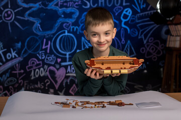 Young boy assembling a wooden model ship with focus and concentration, surrounded by scattered...