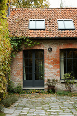 A modern farmhouse-style brick house with black steel windows and terracotta roof tiles, surrounded by an old stone courtyard in Lille, France. The scene includes a small garden with ivy-covered walls