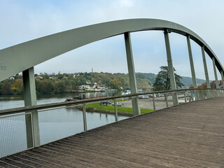 A pedestrian bridge arches gracefully over a calm river, surrounded by trees and hillside homes during a cloudy day