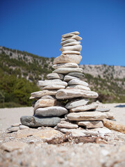 Close-up of a carefully balanced stone stack on a sandy beach, with a rugged landscape and bright blue sky in the background. This scene captures the serenity and beauty of nature.