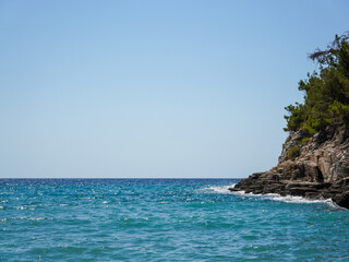 Clear turquoise waters meet a rugged rocky coastline lined with pine trees under a cloudless blue sky, showcasing the peaceful and untouched beauty of a secluded seascape.