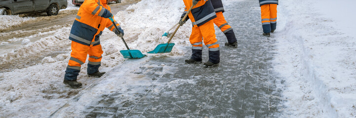 A group of workers in orange and black coats are shoveling snow from a sidewalk