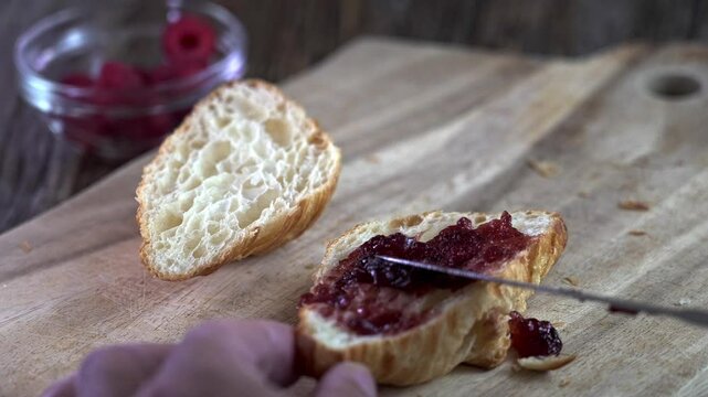 A person carefully spreads rich homemade raspberry jam onto a warm, flaky croissant while sitting at a rustic wooden table. The cozy kitchen adds to the enticing atmosphere.