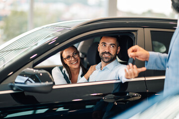 Happy couple inside car receiving keys from a salesperson in a dealership