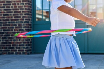 A young girl twirls a colorful hula hoop around her waist, bathed in warm sunlight against a backdrop of a brick wall and turquoise door.