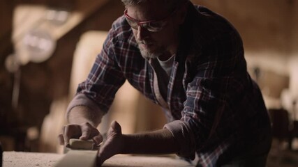 Slow-motion shot of a professional male carpenter sanding wood on the work bench and blowing dusts