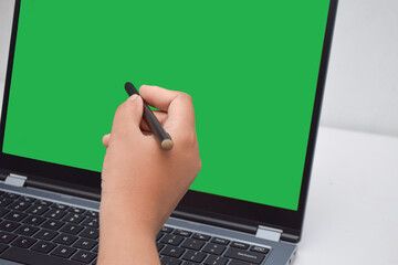 Close-up of a hand holding a stylus, pointing at a blank green screen on a laptop. set up for...