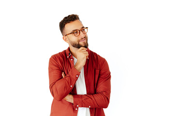 Attractive arabian or caucasian man with glasses, dressed in a red casual shirt with beard, holding his hand on a chin, standing on isolated white background, thinking about something and looking away