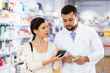 Obraz premium Female shopper shows name of drug on the smartphone screen. Pharmacist helps to find medicine in pharmacy