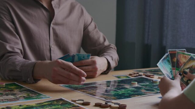 Tilt up shot of young curly-haired man rolling dice, showing card and making move on map while playing tabletop game with girlfriend at home