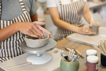 Female hands craftspersons in striped aprons are busy molding clay products. They are sitting near work table, making plate out of raw clay. Working together in pottery workshop.