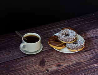 A cup of hot black coffee on a saucer with a spoon and a plate with several donuts in chocolate icing on a wooden dark table.