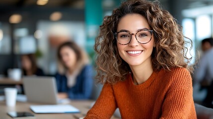 Cheerful young female professional woman with curly hair and glasses wearing an orange sweater sitting at a desk and using a laptop computer in a coworking or office environment