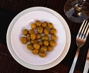 On small square white plate lies portion of large ripe green olives with stone. Snack is supplemented with glass of beer