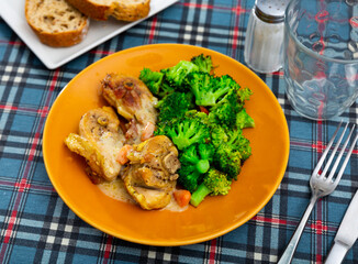 Parts of chicken carcass cooked with sour cream and carrot sauce, garnished with boiled broccoli cabbage. Dish decoration, table setting for lunch, main course and cutlery.