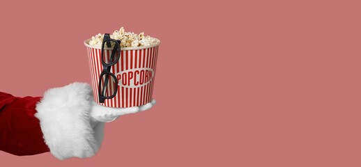 Santa's hand holding bucket of popcorn and 3D glasses on pink background with space for text
