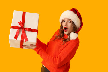 Surprised young woman in Santa hat with Christmas gift box on yellow background