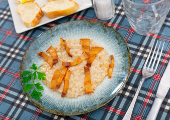 There is portion of boiled oatmeal porridge with pieces of fried bacon on plate. Hearty lunch, complemented by sliced bread, cutlery.