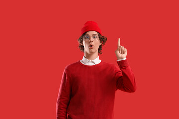 Surprised young man in red clothes pointing up on color background