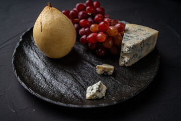 Blue blue cheese Garganzola or Dor Blue with a bunch of red grapes and a yellow pear on a black mica board on a dark background. Space for text. . High quality photo