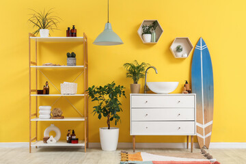 Modern sink, chest of drawers, shelving unit and surfboard near yellow wall in interior of bathroom