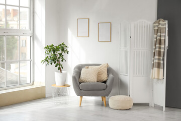 Grey armchair, houseplant, pouf and white folding screen in interior of light living room