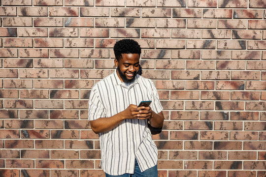 Young cheerful african american guy standing outdoors with cellphone in hands and smiling at it.